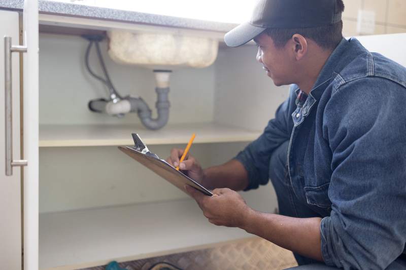 A home inspector makes notes about what he sees with the plumbing under a sink.