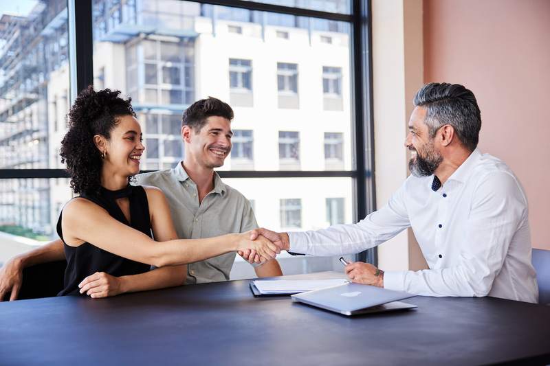 Smiling couple shaking hands with a broker after signing a mortgage loan.