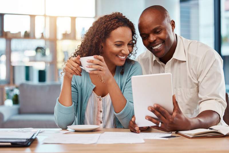 Woman with mug and man smile while reviewing financial information on a tablet.